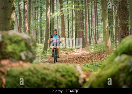 Hübsche ältere Frau unterwegs mit ihrem Elektro-Mountainbike auf einem felsigen Waldweg in der Fränkischen Schweiz, Bayern, Deutschland Stockfoto