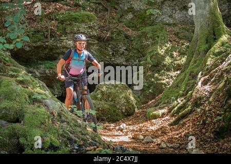 Hübsche ältere Frau unterwegs mit ihrem Elektro-Mountainbike auf einem felsigen Waldweg in der Fränkischen Schweiz, Bayern, Deutschland Stockfoto