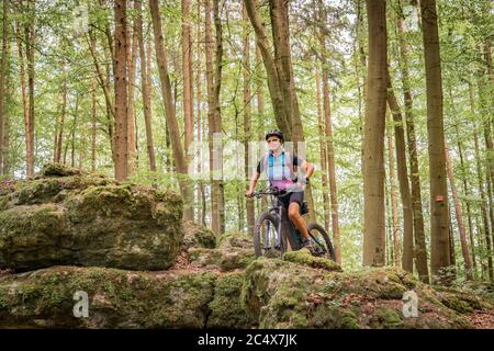 Hübsche ältere Frau unterwegs mit ihrem Elektro-Mountainbike auf einem felsigen Waldweg in der Fränkischen Schweiz, Bayern, Deutschland Stockfoto
