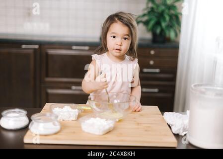 Schöne niedliche kleine Mädchen trägt weißes Kleid ist Whisking Eier in Rührschüssel mit Gabel Stockfoto