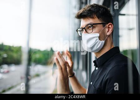 Trauriger Mann mit Gesichtsmaske, der durch Fenster, Quarantäne und Sperrkonzept hinaus schaut. Stockfoto