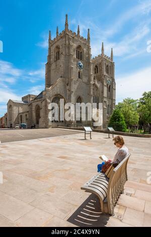 Frau liest Buch Konzept, Blick im Sommer von einer reifen Frau sitzen allein auf einer Bank in der Nähe einer Kathedrale in einer europäischen Stadt, King's Lynn, Großbritannien Stockfoto