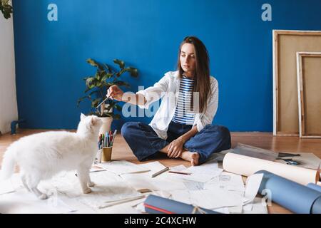 Nachdenkliches Mädchen auf dem Boden mit Zeichnungen sitzen traumhaft Blick auf weiße Katze in der Nähe, während die Zeit zu Hause Stockfoto