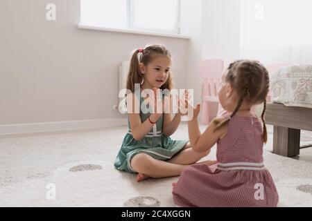 Nette kleine Schwestern spielen zusammen sitzen auf dem Teppich in identischen Kleidern verschiedenen Farben in einem Raum mit viel Licht Stockfoto