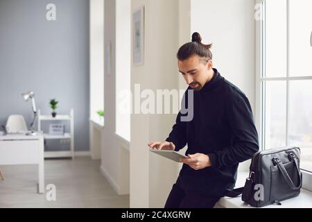 Ein ernster Kerl steht mit einem Tablett in der Hand, während er im Büro am Arbeitsplatz steht. Stockfoto