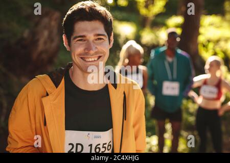 Männlicher Läufer, der draußen steht und die Kamera anschaut und lächelt. Sportler, der nach einem Lauf mit anderen Athleten im Hintergrund eine Pause einlegt. Stockfoto