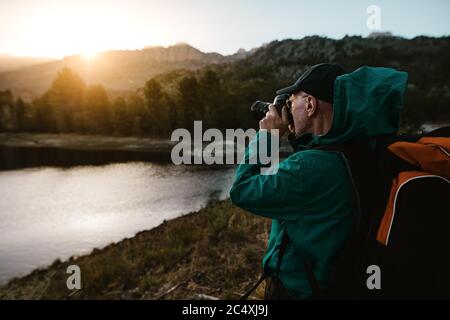 Alter Mann auf Wanderurlaub fotografieren schöne Aussicht mit einer Digitalkamera. Älterer Mann am Fluss im Wald stehen und Fotos machen. Stockfoto