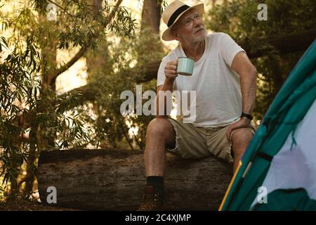 Rentner, der auf einem Holzbuch mit einer Tasse Kaffee sitzt. Ältere männliche Camping im Wald mit einem erfrischenden Kaffee. Stockfoto