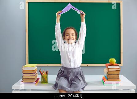 Zurück zur Schule. Fröhliches kleines Mädchen, das auf dem Schreibtisch sitzt und das Buch über ihrem Kopf im Klassenzimmer hält. Nettes Kind Spaß in der Schule Stockfoto