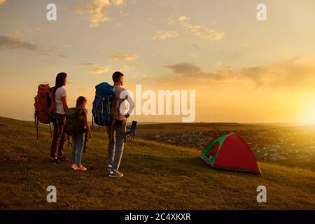 Familiencamping in den Bergen. Eltern und Kind mit Rucksäcken genießen schönen Sonnenuntergang auf dem Campingplatz, Kopierer Platz Stockfoto