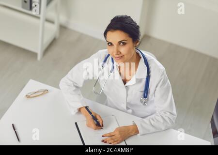 Selbstbewusste Frau Ärztin Kinderärztin schreibt in einer Zwischenablage, die an einem Tisch in einem weißen Büro des Krankenhauses sitzt Stockfoto