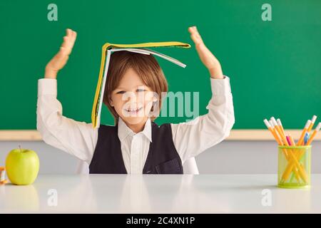 Zurück zur Schule. Fröhlicher kleiner Junge versteckt unter Buch, spielen dummes Spiel am Schreibtisch im Klassenzimmer. Stockfoto