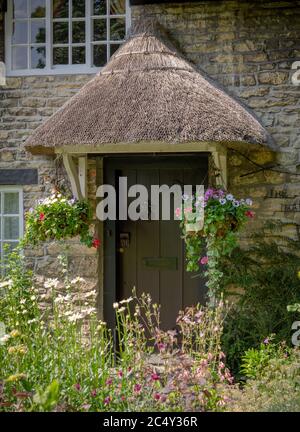 Strohgedeckte Tür eines malerischen Häuschens in Thornton-le-Dale, North Yorkshire mit zwei hängenden Körben auf jeder Seite. Blumen stehen im Vordergrund. Stockfoto