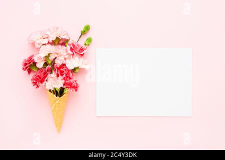 Waffel Eis-Kegel mit roten und weißen Nelkenblumen und leeres Papier Blatt auf rosa Hintergrund. Modell, flach liegend. Sommerkonzept. Kopierbereich oben Stockfoto