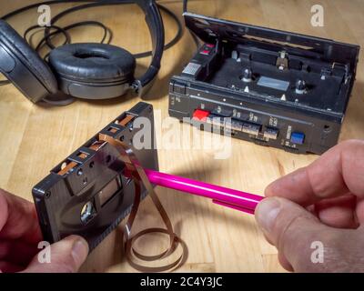 Die Hände eines Mannes halten eine Audiokassette ohne Marke, mit einem rosa Stift in lose Kassette zu Spule, mit einem persönlichen Kassettenspieler und Kopfhörer hinter. Stockfoto