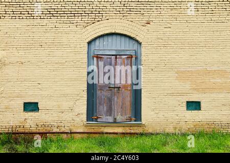 Ein lackiertes gelbes Ziegelgebäude mit vertaufter grüner Eingangstür und Grasfront mit rostigen Rohren Stockfoto