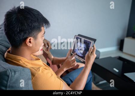 Asiatische schöne schwangere Frau und Mann suchen Ultraschall-Scan auf einer Couch im Wohnzimmer zu Hause. Konzept der Schwangerschaft Gesundheitsversorgung. Stockfoto