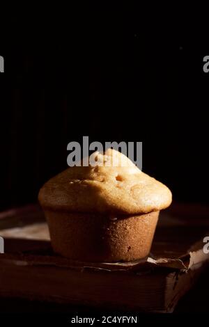 Hausgemachter Muffin Cupcake liegt auf altem Buch auf schwarzem Hintergrund Stockfoto