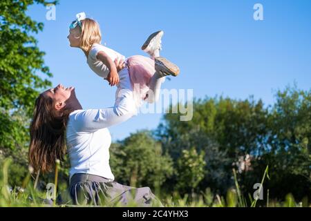 Mama hebt Tochter hoch in blauen Himmel. Glückliche Mutter und Mädchen spielen Spaß im Park im Sommer zusammen auf dem grünen Gras. Familienleben. Stockfoto