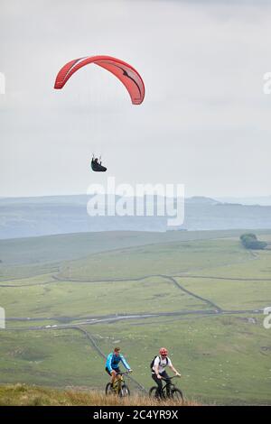 Paragliding vor dem Mam Tor Hügel in der Nähe von Castleton in der High Peak of Derbyshire, England, während Radfahrer entlang der Spitze fahren Stockfoto