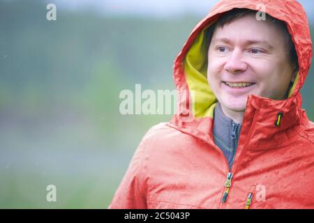 Ein junger Mann im Regenmantel mit Kapuze steht im Regen. Regentropfen fallen auf Gesicht und Jacke. Stockfoto