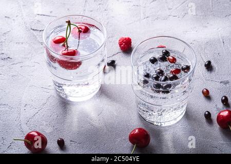 Frisches kaltes Mineralwasser trinken mit Kirsche, Himbeere und Johannisbeere Beeren in zwei transparenten Glas auf Stein Beton Hintergrund, Sommer Diät beverag Stockfoto