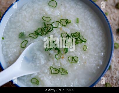 Einfacher Reis Congee, Reisbrei, mit einem Spritzer gehackter Frühlingszwiebel darauf serviert in einem chinesischen Bogen mit Löffel. Stockfoto