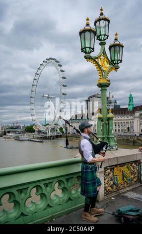 Ein Dudelsack-Spieler in schottischer Hochlandkleidung, der auf der Westminster Bridge mit dem London Eye und der Themse im Hintergrund unterwegs ist. Stockfoto