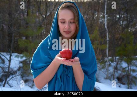 Ein Mädchen in einer blauen Kapuze mit einem roten Apfel in der Hand.das Mädchen schaut auf den Apfel in der Hand.eine Frau im Winterwald zu Weihnachten Stockfoto