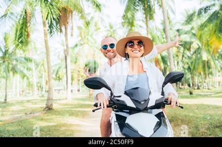 Glücklich lächelnd Paar Reisende Reiten Motorrad Roller unter Palmen. Lachender Mann, der eine Kokosnuss in der Hand trägt.Tropisches Urlaubskonzept Stockfoto