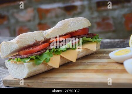 Ein appetitliches Sandwich mit Schinken, Käse, Tomaten und Salat liegt auf einem Brett mit Ei und Zitrone, auf einem Holztisch, handwerklich Stockfoto