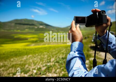 Mann mit Kamera, der ein Panoramabild in Richtung Castelluccio di Norcia (Juni 2020) macht, mit Feldern in üppiger Farbe für den Beginn der Blüte. Stockfoto