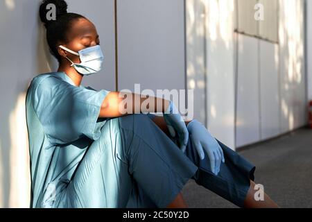 Müde erschöpft Frau african Krankenschwester tragen Gesichtsmaske Handschuhe sitzen auf Krankenhausboden. Stockfoto