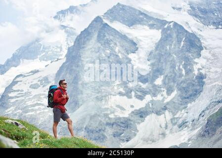 Horizontale Aufnahme eines riesigen verschneiten Berges in Schweizer Alpen, der den ganzen Hintergrund nimmt, männlicher Wanderer mit einem Rucksack, der auf grüner Wiese im Vordergrund steht. Konzept des Tourismus, Wandern, Trekking. Stockfoto