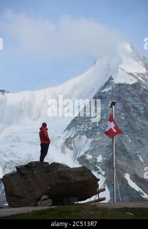 Männliche Touristen mit Blick auf schöne Berglandschaft. Mann auf Steinen in der Nähe der Fahnenstange mit Schweizer Flagge. Ober Gabelhorn ist im Hintergrund. Wilde Natur mit herrlichem Ausblick. Tourismus in den Alpen. Stockfoto