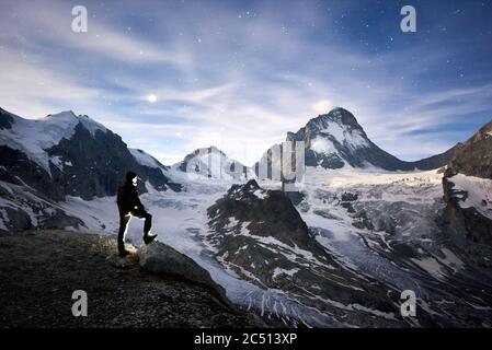 Horizontale Momentaufnahme des unglaublichen blauen Himmels voller Sterne und des Mondes über kühlen ewigen schneebedeckten Gipfeln der Pennine Alpen in der Schweiz, ein touristisches Standing und die Schönheit Dent Blanche beobachten Stockfoto