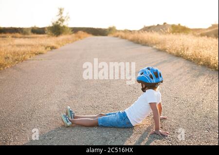 Müde kleines Kind trägt Sportausrüstung Helm und Turnschuhe sitzen auf leeren Asphaltstraße im Sommer warmen Sonnenuntergang Stockfoto