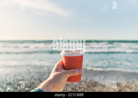 Ein junger Mann hält eine Papiertasse mit Kaffee auf dem Hintergrund des Meeres Stockfoto