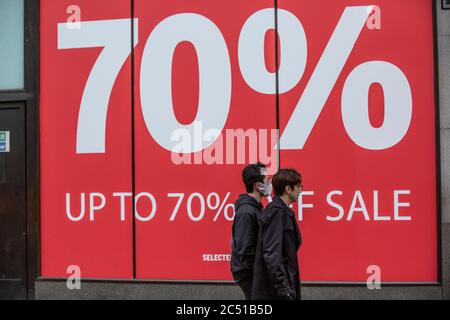 Käufer mit Gesichtsmasken kommen an einem großen Schaufenster vorbei, das eine 70%ige Verkaufsermäßigung auf der Oxford Street, West End, London, Großbritannien, anwirbt Stockfoto