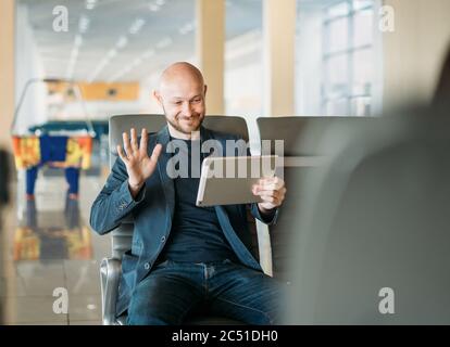 Schöner kahlbärtiger lächelnder Mann Geschäftsmann im Anzug kommunizieren mit jemandem von Tablet in der Flughafenlounge Stockfoto