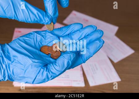 Kleine Metallmünzen in menschlicher Hand und Geldbestellungen auf Holzschreibtisch. Euro Bargeld auf Handfläche in blauem Schutzhandschuh zählen. Rechnungen Zahlung in COVID-19 Pandemie. Stockfoto