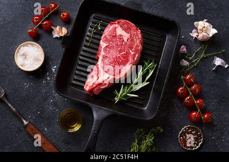 Draufsicht Black Angus Prime Beef Rib Eye Steak auf Gusseisen Grill Pfanne mit frischem Rosmarin, Kirschtomaten, Olivenöl und Gewürze. Kreatives Layout w Stockfoto