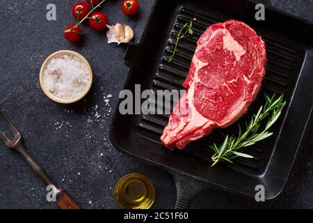 Draufsicht Black Angus Prime Beef Rib Eye Steak auf Gusseisen Grill Pfanne mit frischem Rosmarin, Kirschtomaten, Olivenöl und Gewürze. Kreatives Layout w Stockfoto