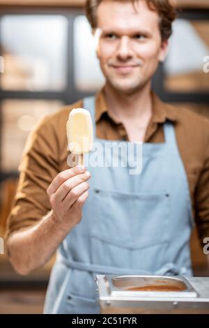 Portrait eines hübschen Verkäufers in Uniform mit weißem Schokoladeneis auf einem Stock im Laden Stockfoto