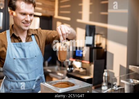 Der Verkäufer taucht Eis in die heiße geschmolzene Schokolade und macht Eis auf einem Stock mit Belag Stockfoto