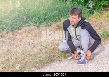 Junger Mann, der sich bereit ist, an sonnigen Tagen draußen zu laufen. Welpe hilft, Jogging Schuhe von Kerl binden. Männlich Biegen über binden seine Schnürsenkel auf seinem Laufschuh Stockfoto