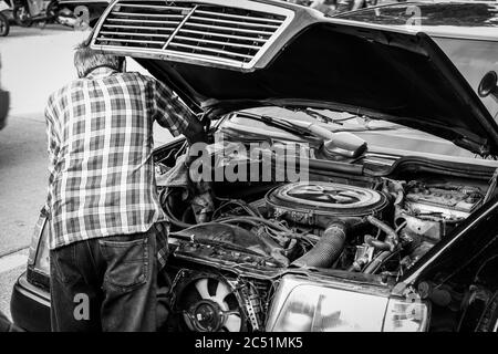 Sie sehen ein schwarz-weißes Bild eines alten Autos auf einer Straße mit geöffneter Motorhaube und einem von einem Menschen reparierten Motorraum Stockfoto