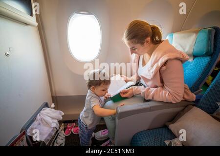 Eine junge schöne Mutter sitzt in einem Flugzeugsessel und zeigt ihrem kleinen niedlichen Baby, das vor ihr steht ein Buch. Nahaufnahme, Weichfokus, oben Stockfoto