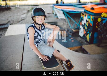 Junge sitzt auf Skatepark, Kamera schauen. Kind ruht mit Skateboard im Skatepark. Kind sitzt auf Rampe ruht nach dem Training auf Skateboard. Trendy Stockfoto