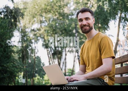 Bärtiger Mann mit Laptop-Computer, online einkaufen, auf Bank sitzen. Erfolgreicher Freiberufler, der im Park arbeitet, auf der Tastatur tippt, auf die Kamera schaut Stockfoto